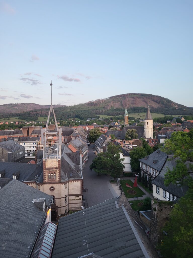 Luftaufnahme einer kleinen historischen Stadt mit engen Kopfsteinpflasterstraßen und grauen Schieferdächern. In der Bildmitte stehen zwei hohe Kirchtürme, umgeben von alten Gebäuden und Bäumen. Im Hintergrund erstrecken sich grüne Felder und ein bewaldeter Hügel unter einem klaren, hellblauen Abendhimmel